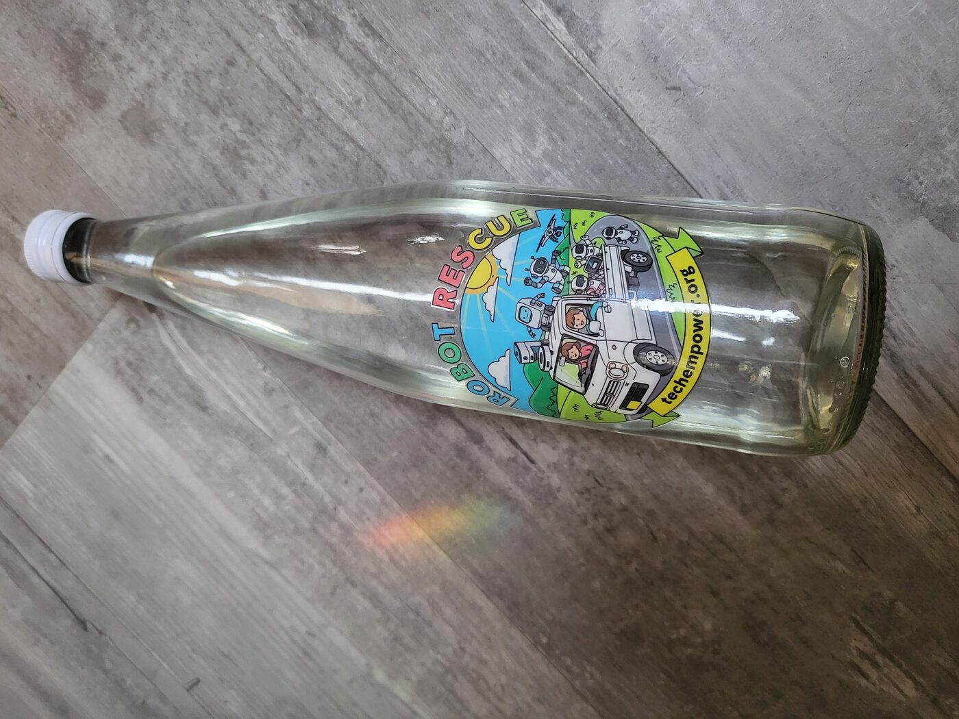 A clear glass bottle with a white cap, lying on a grey wooden floor, with the colourful ROBOT RESCUE decal on its side.